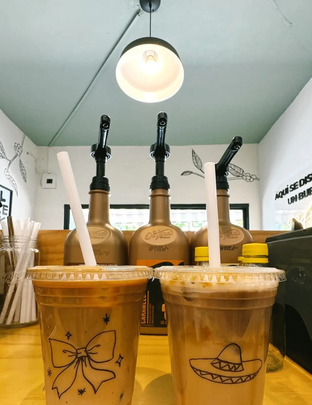 La barra de Sublime con sus jarabes y dos lattes a las rocas con moño y sombrero dibujados en el vaso
